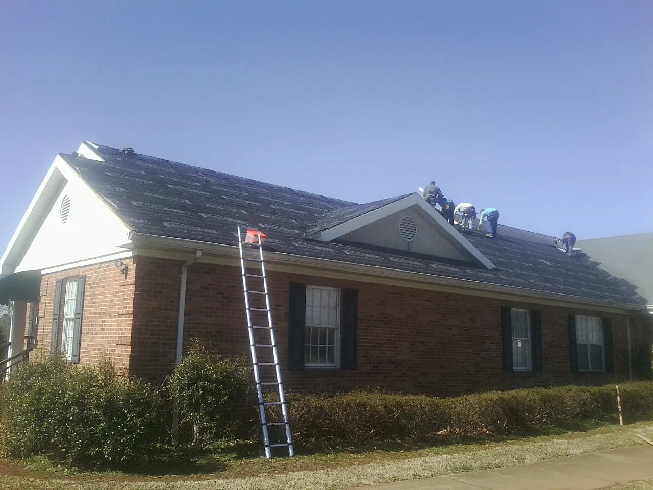 Crew tearing shingles off a brick ranch roof with a ladder set — Emergency Roof Repair work by our crew in Dumont