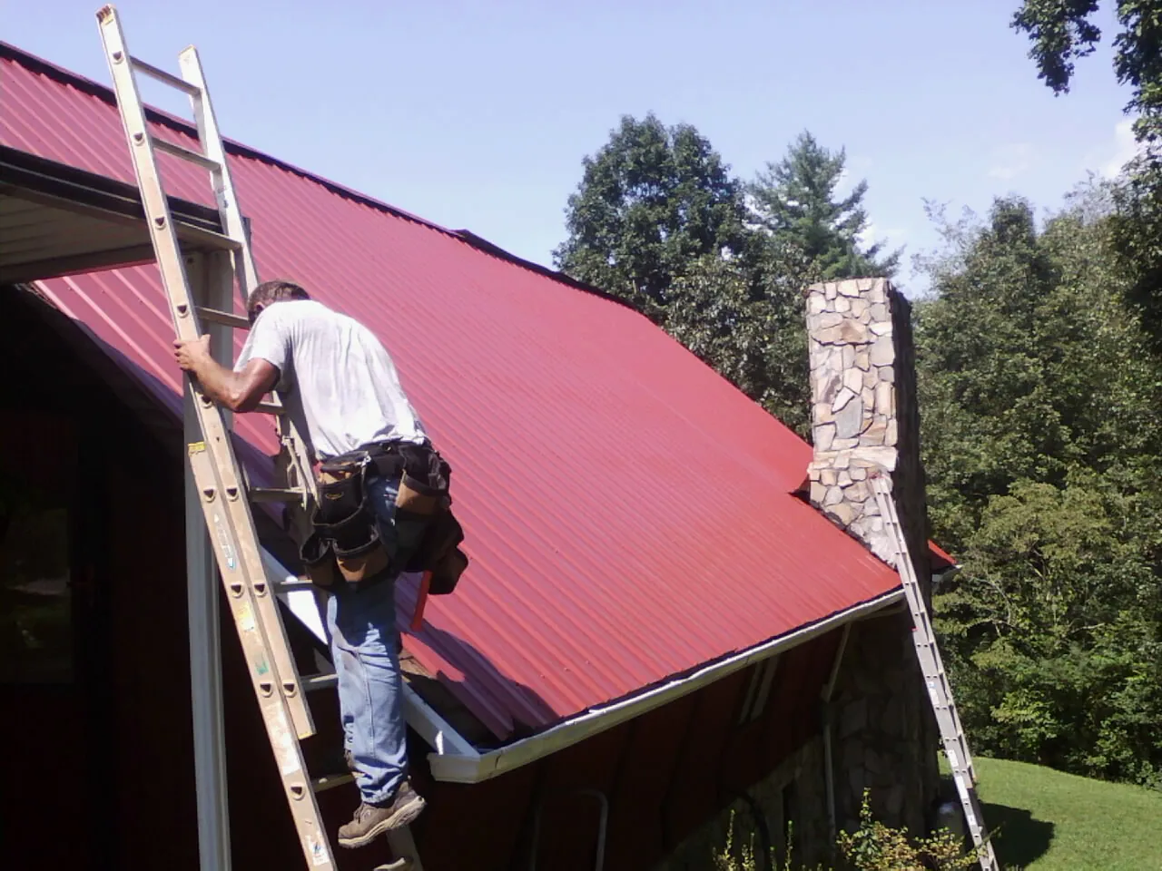 Roofer on a ladder beside a red metal roof and stone chimney — Soffit & Fascia Repair by Roofing Brothers in Dumont